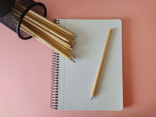 A top view of colored wooden pencils in a pen holder and notepad on the pink surface