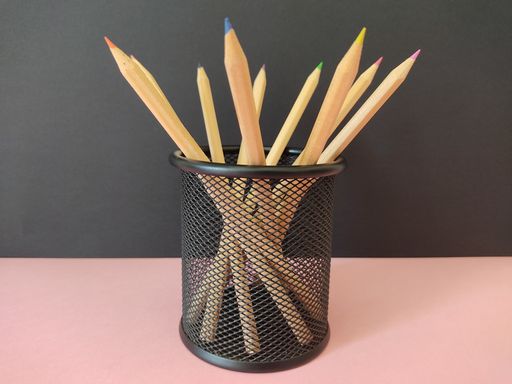Wooden colored pencils in a mesh pen holder on the pink surface in the black background