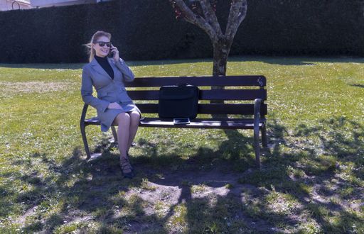 An adult woman wearing a skirt with a blazer and sitting on a bench in a park while talking on the phone
