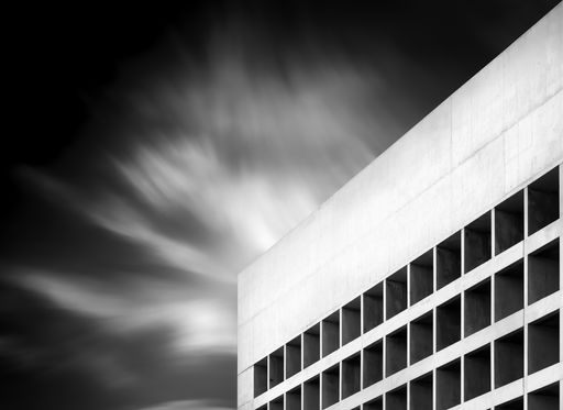 A grayscale shot of a modern building under a cloudy sky with long exposure