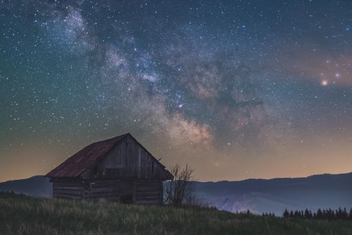 A tiny rural wooden house at the top of a mountain under a night starry sky with Milky Way