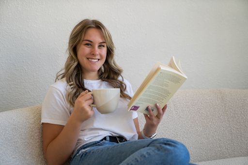 A young woman with a pleasant expression is relaxing on a comfortable couch while perusing a book and sipping a cup of coffee or tea