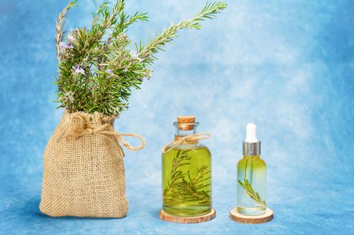 glass bottle and dropper with rosemary essential oil with fresh branches inside next to a raffia bag with freshly cut flowering rosemary branches