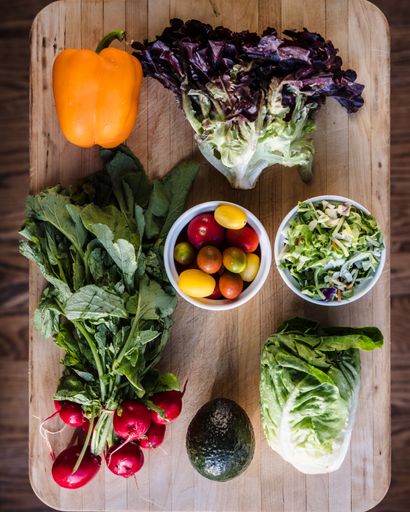 A wooden table displays a cutting board filled with fresh vegetables, including tomatoes, peppers, onions, and carrots