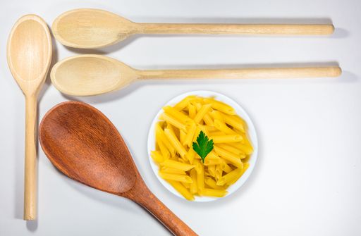 A closeup and top view of wooden spoons and a pot with raw pasta on a white background.