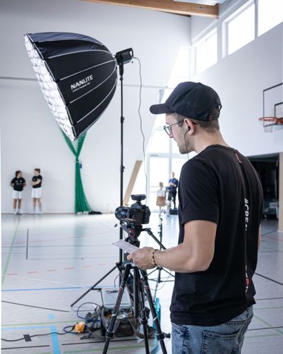 A mature man stands confidently holding a recording device next to a professional camera, poised to capture an important moment