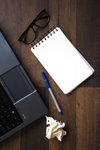 A top view of a wooden desk with a notebook, a pen, and eyeglasses next to a laptop computer.