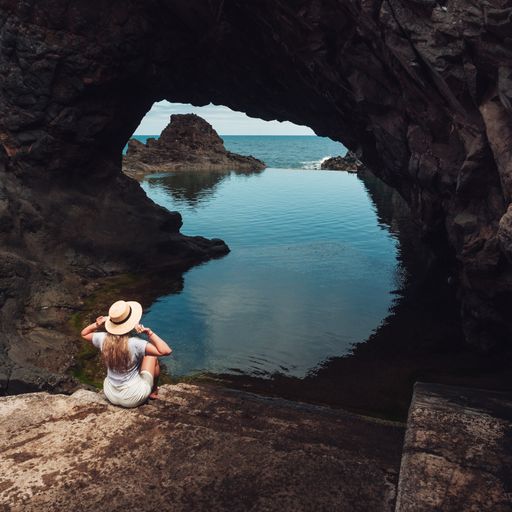 A vertical shot of a young lady sitting on a rock enjoying the view of the calm sea through a cave