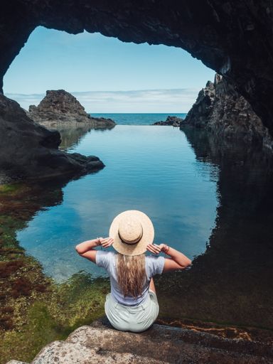 A vertical shot of a young lady sitting on a rock enjoying the view of the calm sea through a cave