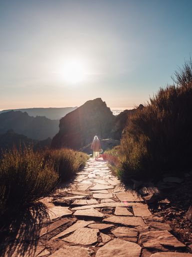 A woman walking on a narrow stone footpath on the hill with mountains in the background at sunset