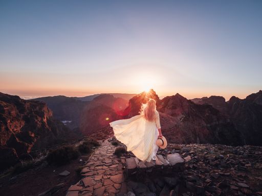 A female from a back in a white dress holding a hat in her hand in a rocky area at sunset