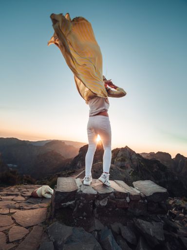 A female standing on the stone wearing white trousers and a yellow jacket waving by the wind in the air