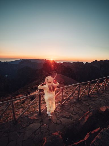 A vertical shot of a female standing near the wooden fence in a mountainous area looking at the sunset