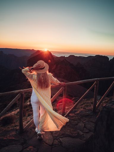 A vertical shot of a female standing near the wooden fence in a mountainous area looking at the sunset