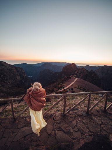 A back view of a woman admiring nature while standing by the fence at the top of a mountain at sunset