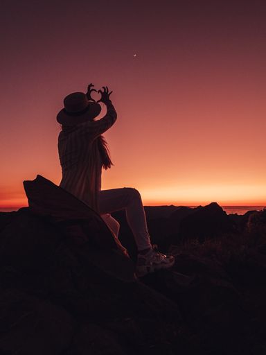 A silhouette of a woman with a hat making a heart with her hands against red sunset sky