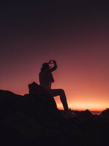 A vertical silhouette shot of a woman out in nature during sunset, her hands shaping a heart