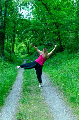 A vertical shot of an excited female with raised arms and a leg on a pathway in a green forest