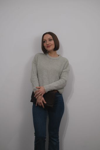 A young adult is standing on a chair indoors, wearing a stylish, comfortable sweater and dark denim jeans