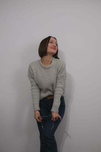 A Caucasian female in her mid-20s is standing with her back against a white wall while her right hand rests on a black purse that is hanging from her