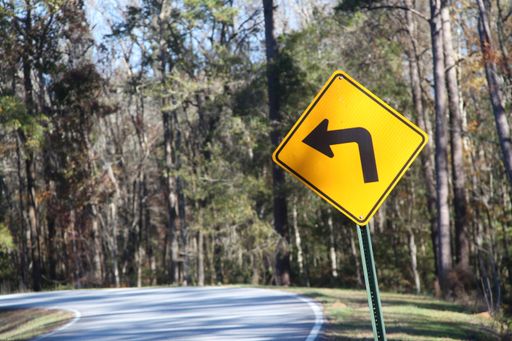 A yellow road sign of the left turn on a road