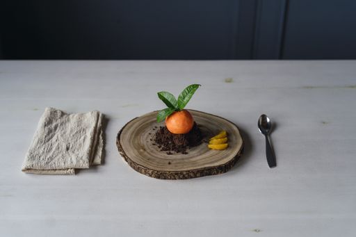 A mandarin shaped pastry on chocolate biscuit crumbles on a wooden board on the table with a spoon and a towel on it