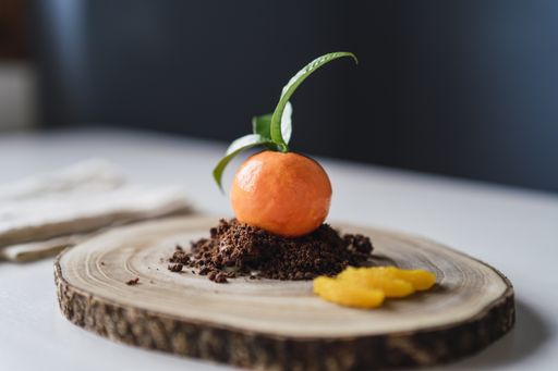 A closeup of a mandarin shaped pastry on chocolate biscuit crumbles on a wooden board