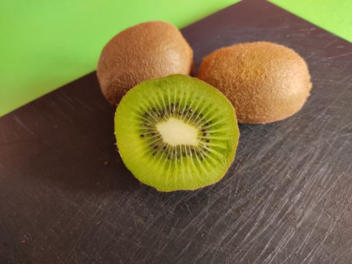 A closeup of a half slice of kiwi on a scratched black cutting board with two whole kiwis behind it