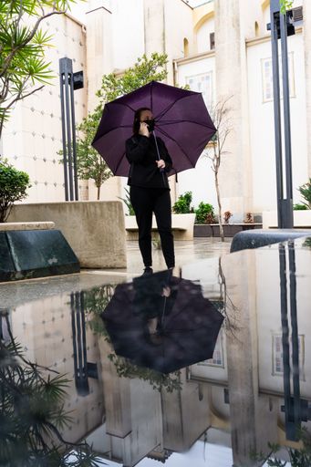 A vertical shot of a female in a black protective mask with a purple umbrella and talking on her phone