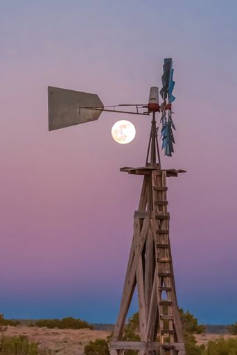 A vertical shot of an old windmill with the full moon in the colorful sky in the background