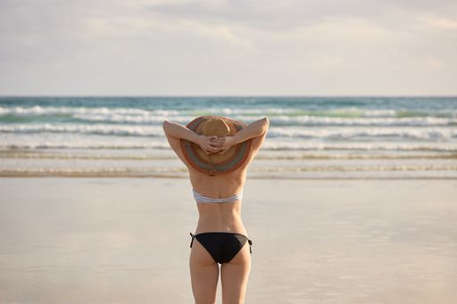 A girl in a bikini and a straw hat walking on the shore of a beach