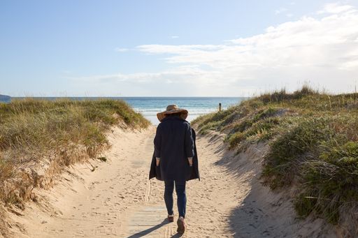 A girl with a straw hat and a black coat walking on a path to the beach
