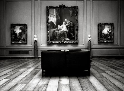 A grayscale interior shot of a museum with a wooden floor, a chair, and a wall with paintings.