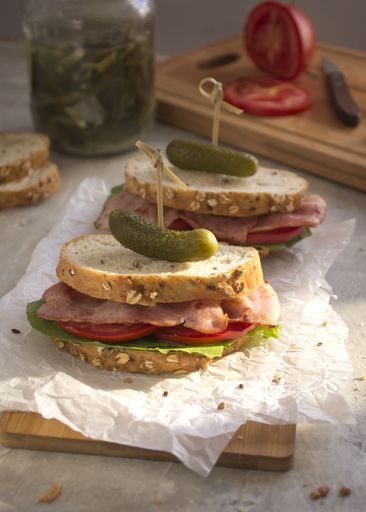 Two bacon sandwiches unwrapped on a wooden board. We can see the tomato and the pickle jar in the background