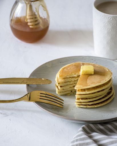 A picture of a stack of pancakes been cut, showing the layers. we can see milk coffee and honey in the background.