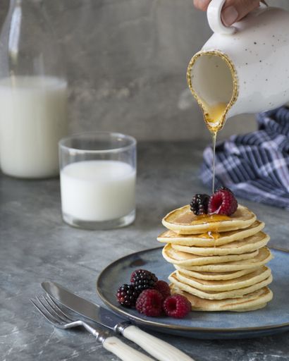 Honey is being poured on a stack of pancakes. We see berries on and on the side of pancakes. A glass of milk and a bottle of milk in the background.