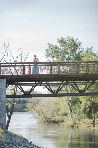 A woman wearing a long dress and standing on a bridge over a river in a park