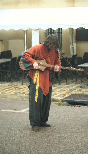 A young man in casual clothing is playing an acoustic guitar on a city street near outdoor tables and chairs