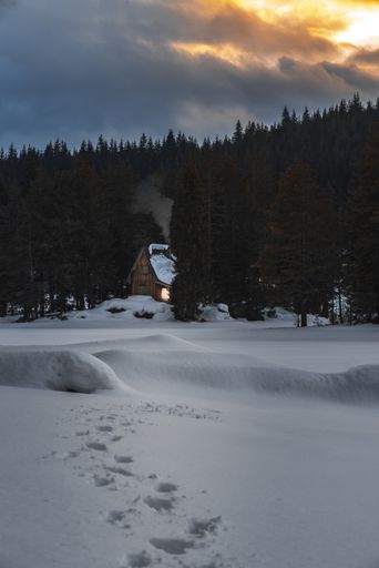 A vertical shot of a small mountain house in the forest with a frozen lake in front at sunset