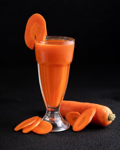 A closeup of freshly cut orange carrots and a glass of carrot juice on a black surface.