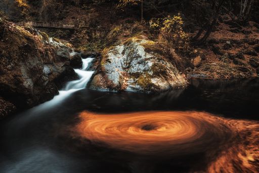 An autumnal scenery of a whirlpool of leaves in the river with a small waterfall in the background
