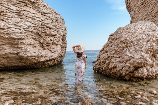 Woman in elegant long white summer dress standing on beautiful Beritnica beach with large rock formations on Pag island in Croatia.