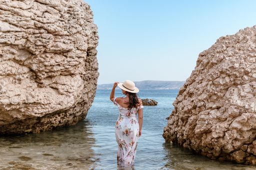 Woman in elegant long white summer dress standing on beautiful Beritnica beach with large rock formations on Pag island in Croatia.