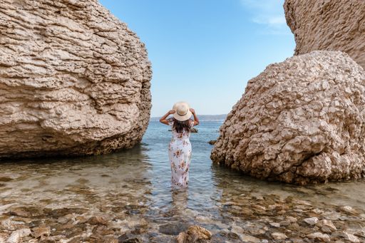 Woman in elegant long white summer dress standing on beautiful Beritnica beach with large rock formations on Pag island in Croatia.