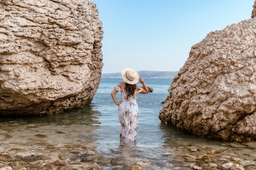 Woman in elegant long white summer dress standing on beautiful Beritnica beach with large rock formations on Pag island in Croatia.