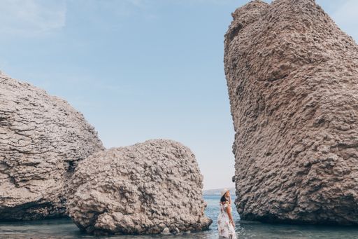 Woman in elegant long white summer dress standing on beautiful Beritnica beach with large rock formations on Pag island in Croatia.
