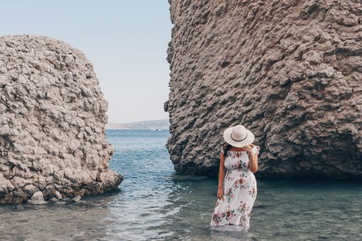 Woman in elegant long white summer dress standing on beautiful Beritnica beach with large rock formations on Pag island in Croatia.