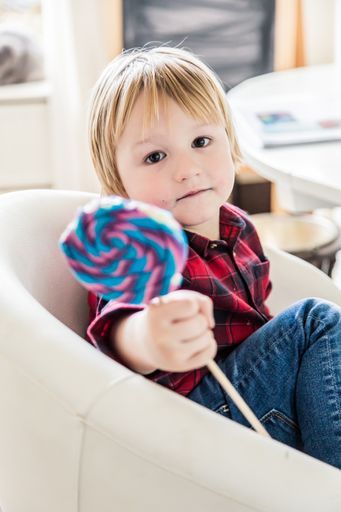 A vertical shot of a blonde Caucasian male toddler indoors with a big candy in his hand