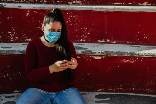 A young caucasian female in a blue medical mask sitting outdoors and texting on her phone