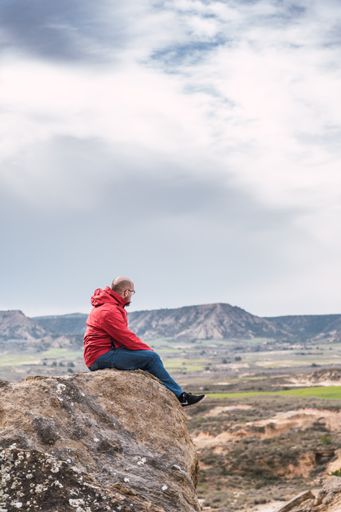 A vertical shot of a male in a red coat sitting on the rock in the hills